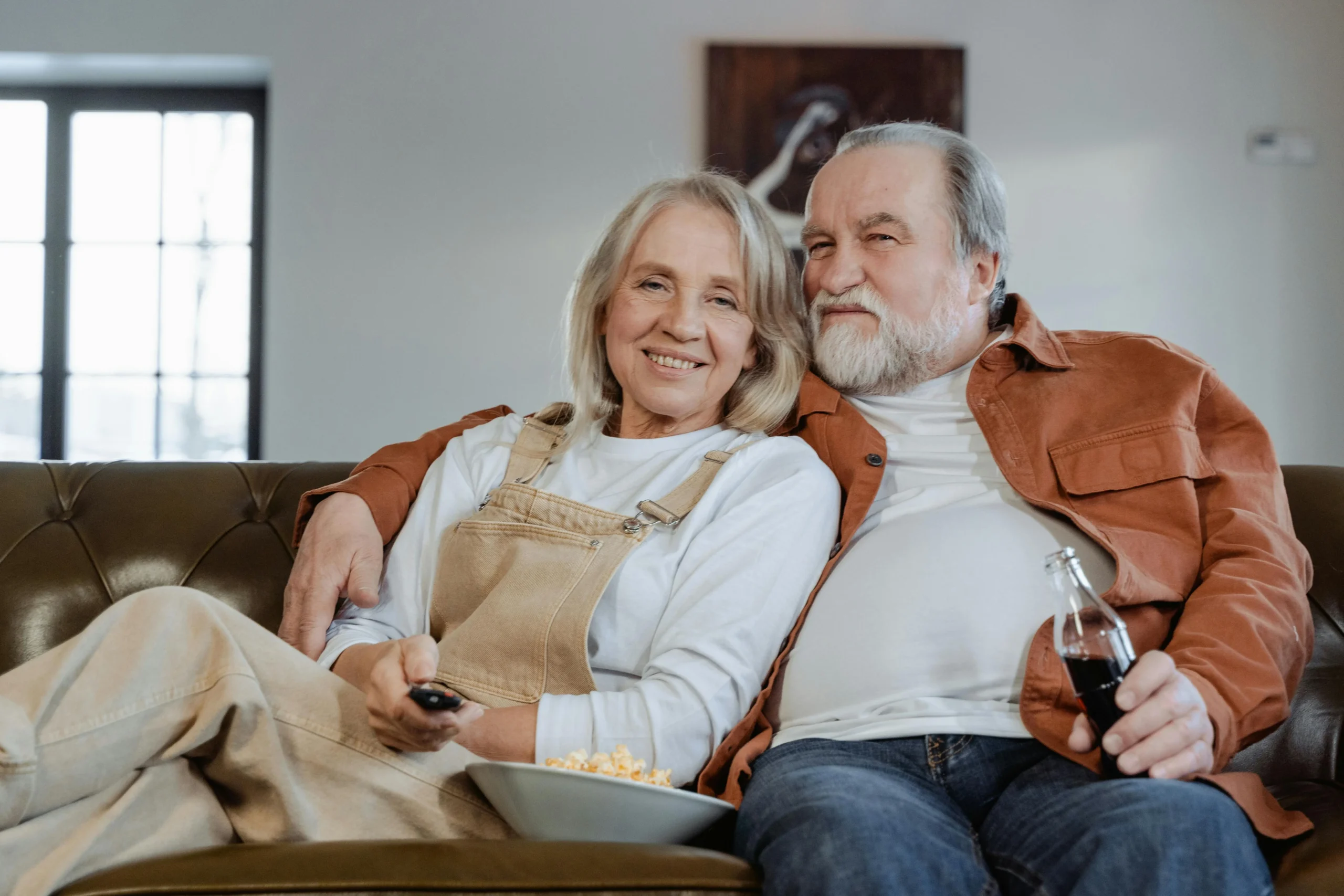Couple de seniors souriant sur un canapé, télécommande et bol de pop-corn devant la télévision